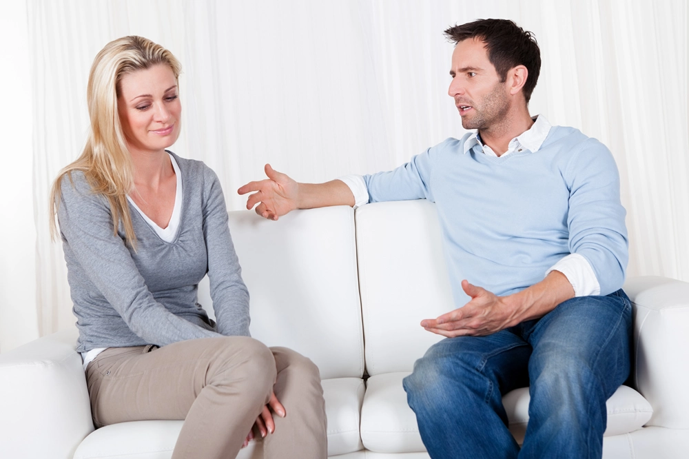 Couple sitting on a couch having a serious conversation, with the man gesturing and the woman looking down