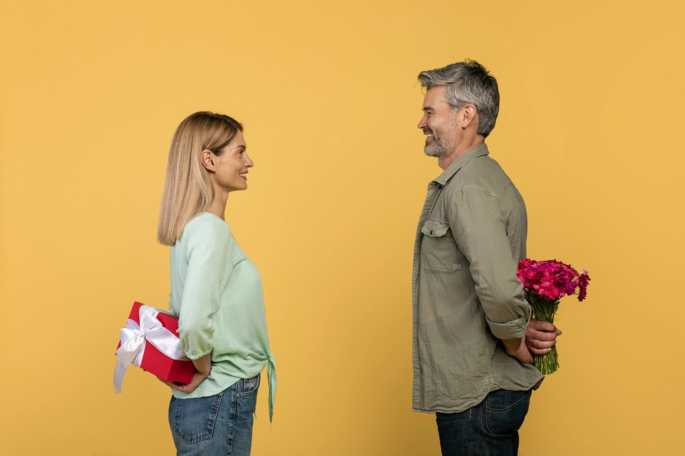 Smiling couple facing each other, hiding gifts behind their backs, symbolizing love and surprise